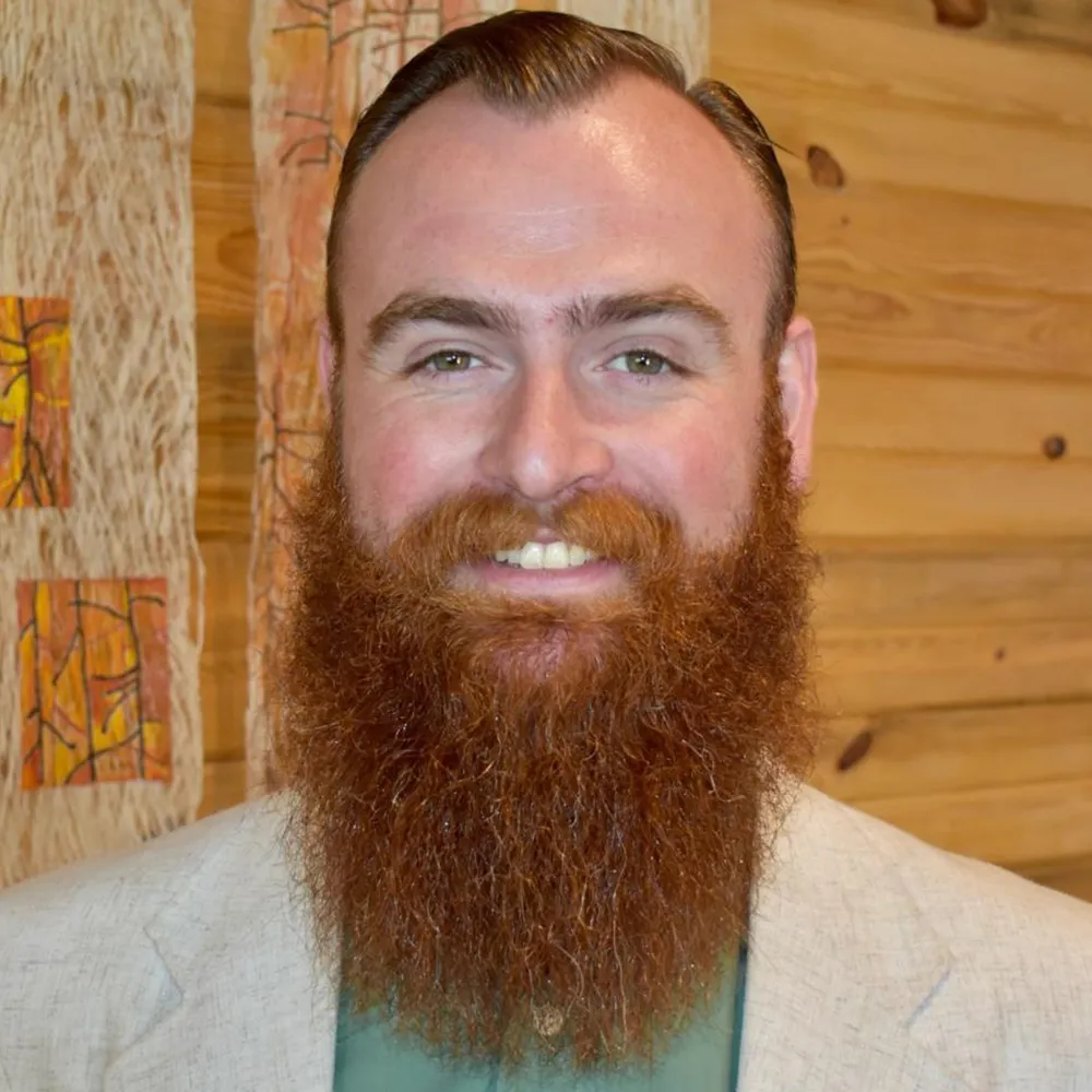 Austin Timpson, a bearded man in a light jacket, smiling against a warm wood-slat wall.
