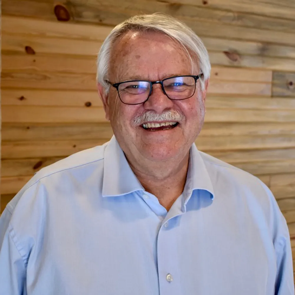 Ben Martin, silver-haired and in glasses, smiling broadly against a warm wood-slat wall.