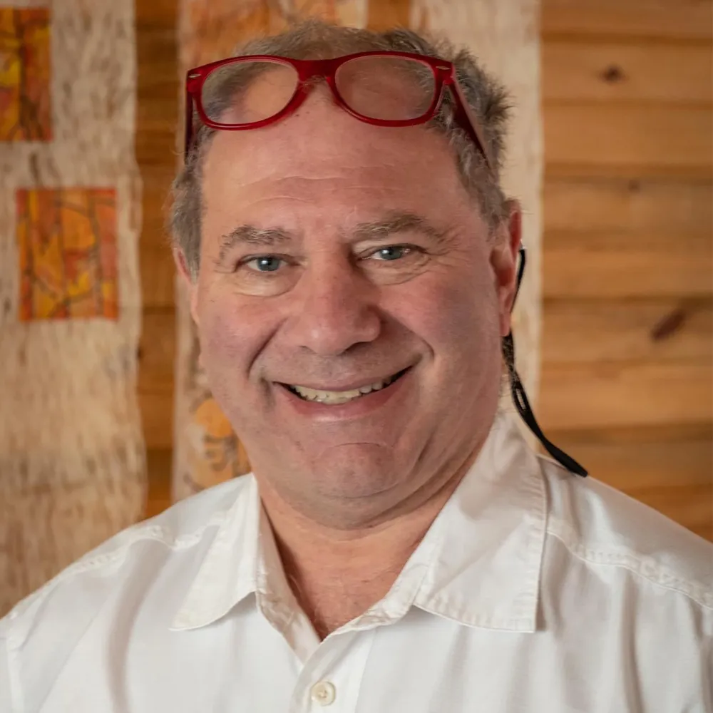 Dan Ziskin smiling, reading glasses pushed up on his head, against a warm wood-slat wall.