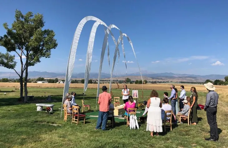 A laying ceremony in an open field, family gathered beneath tall white prayer flags