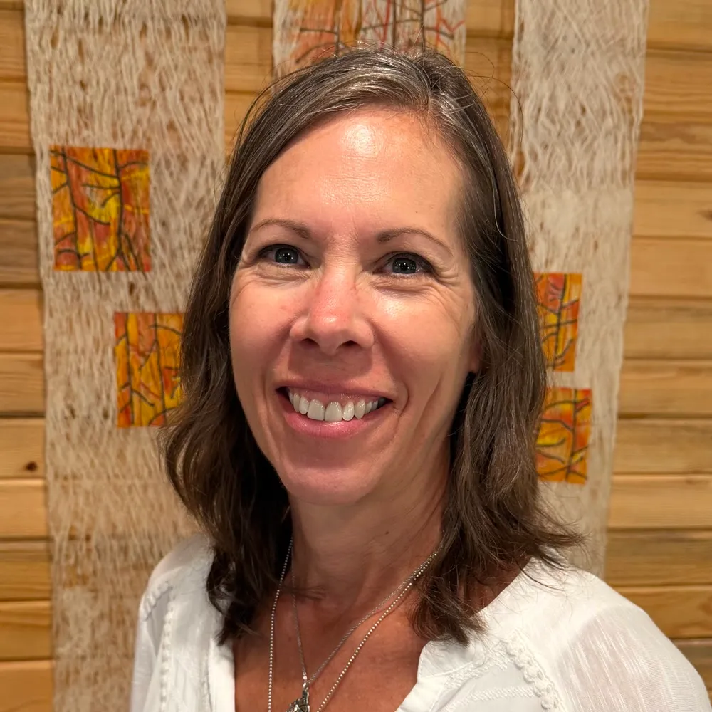 Gretchen Ford smiling in a white embroidered top and pendant necklace, against a warm wood-slat wall.