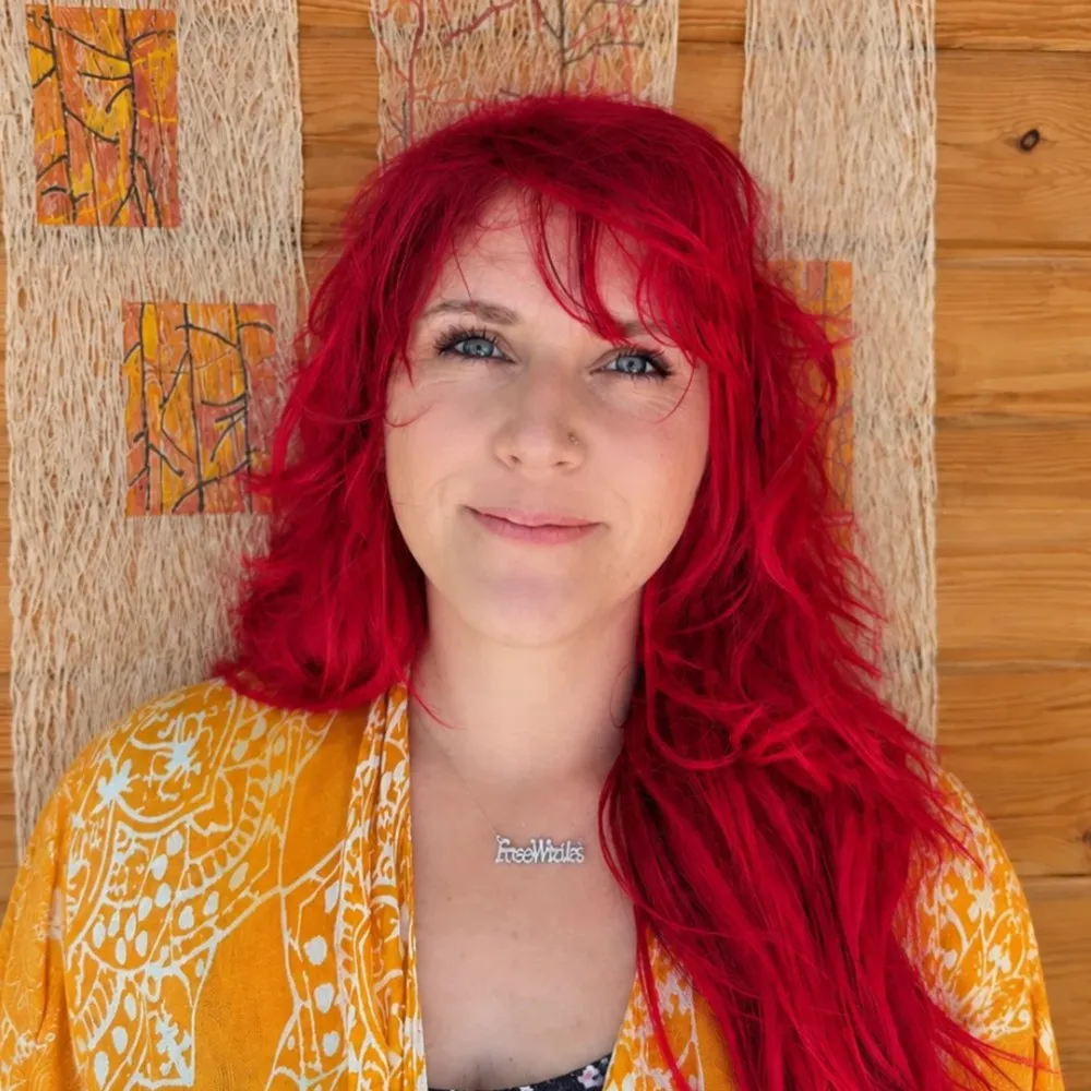 Jen Nenoff with long red hair, smiling softly in a marigold patterned wrap, against a warm wood-slat wall.