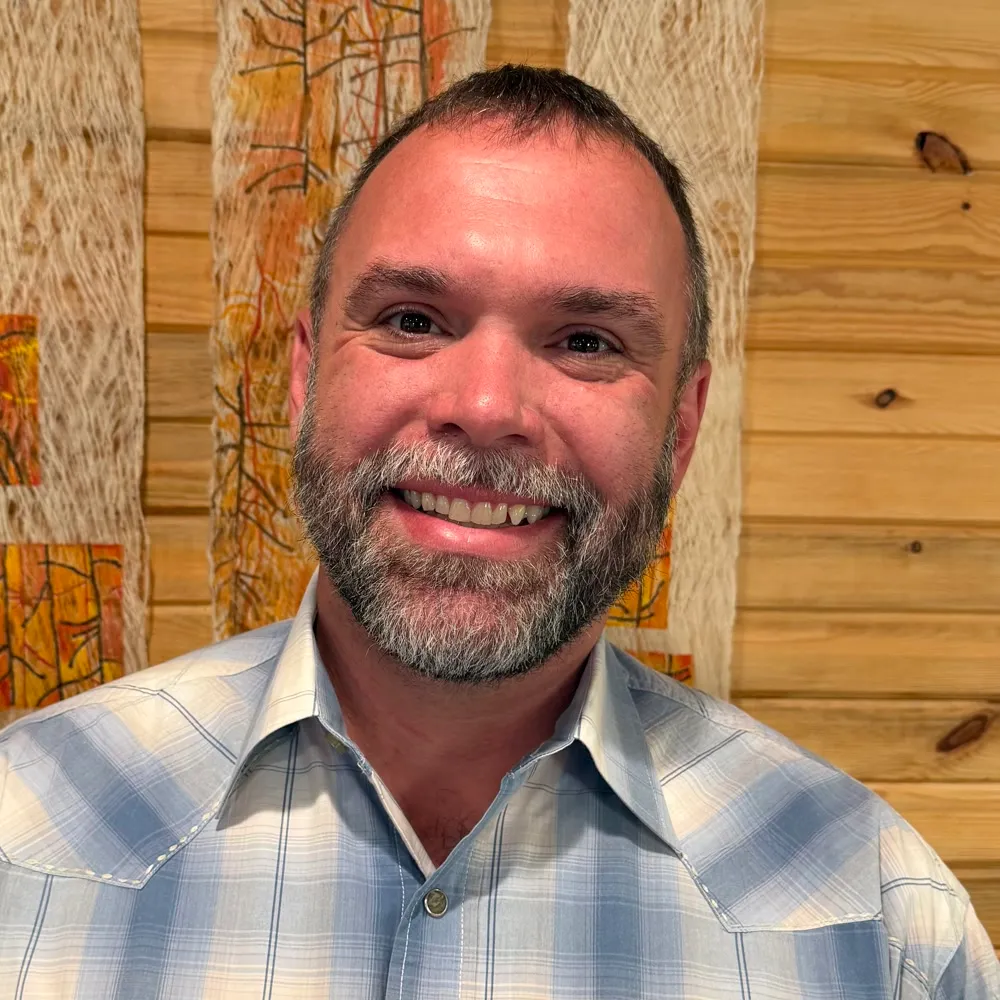 John Knaus smiling in a blue plaid Western shirt, against a warm wood-slat wall.