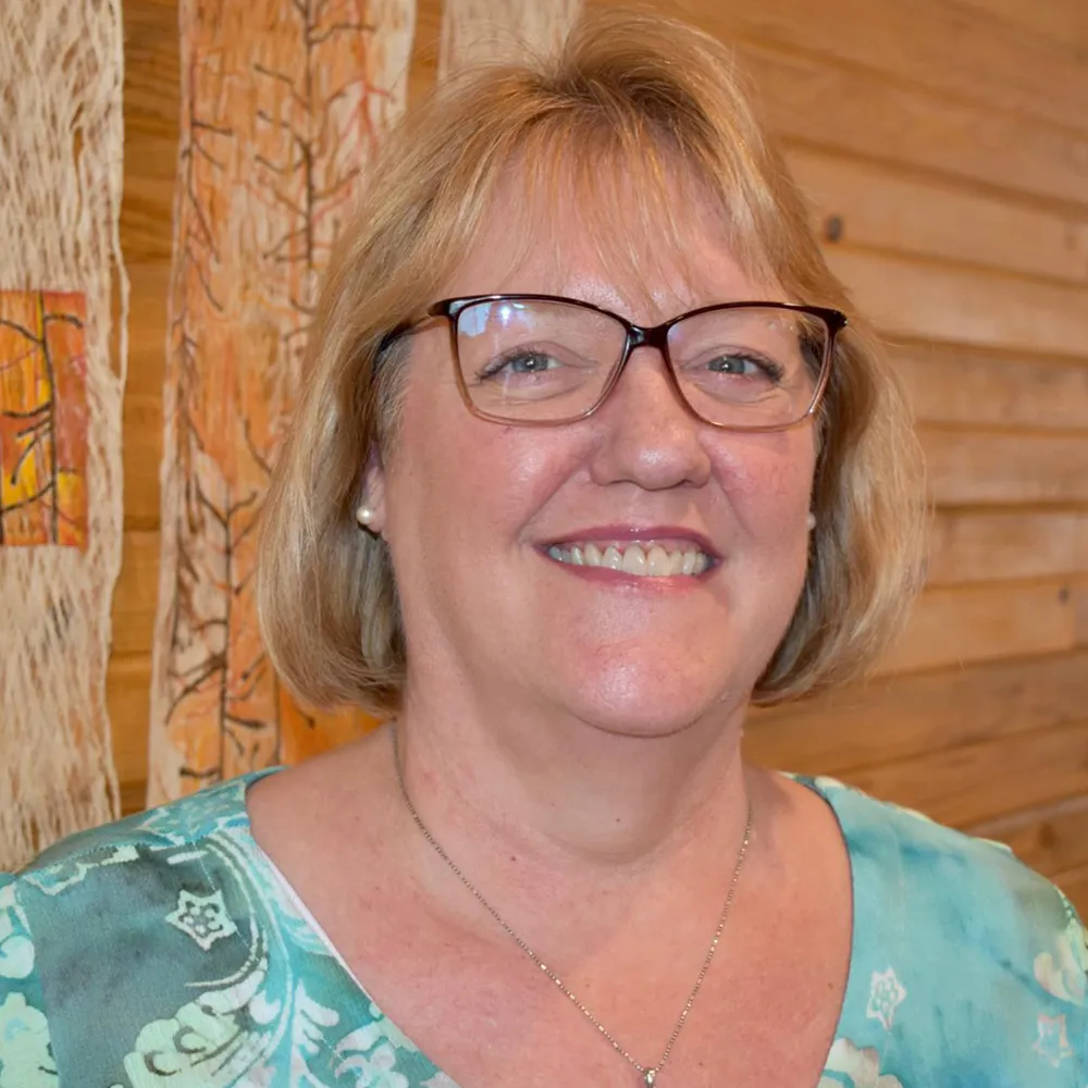 Karis Meskimen smiling in glasses and a patterned teal top, against a warm wood-slat wall.