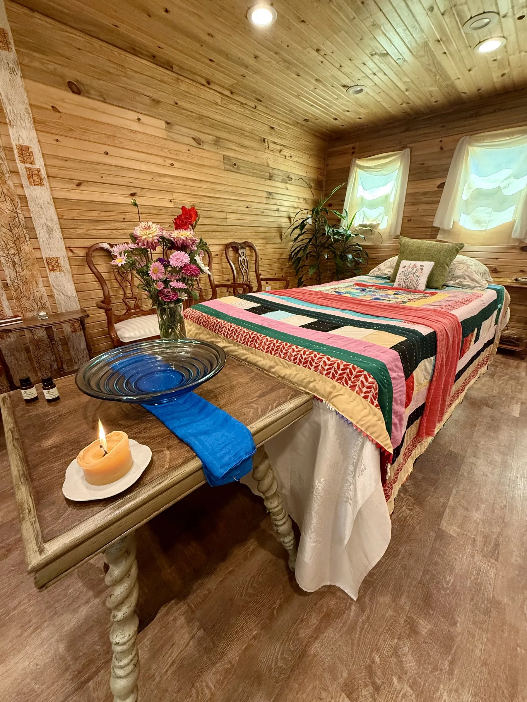 A lit beeswax candle, a blue glass bowl, and a vase of pink and red flowers on a wooden altar table beside the quilt-covered bed in our Lafayette laying-in room