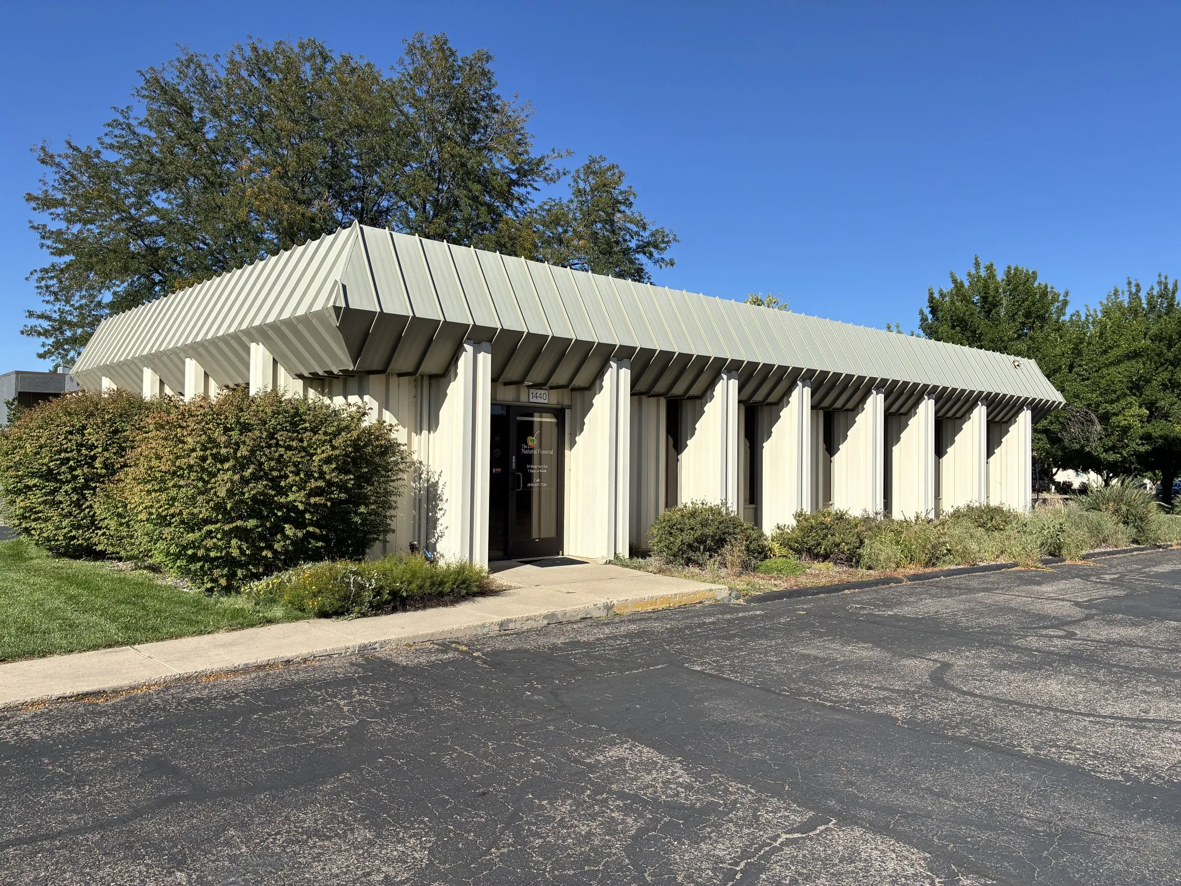 The Natural Funeral's Loveland funeral home at 1440 N Boise Ave, a single-storey building with a ribbed metal roof, shrubs along the front, and trees behind
