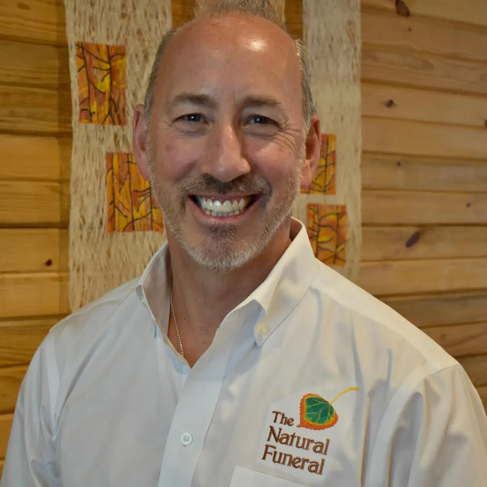 Mike Reagan smiling, wearing a shirt embroidered with The Natural Funeral logo, against a warm wood-slat wall.