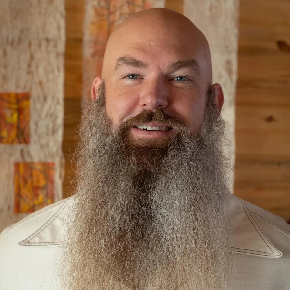 Seth Viddal with a long silver beard, smiling gently, against a warm wood-slat wall.