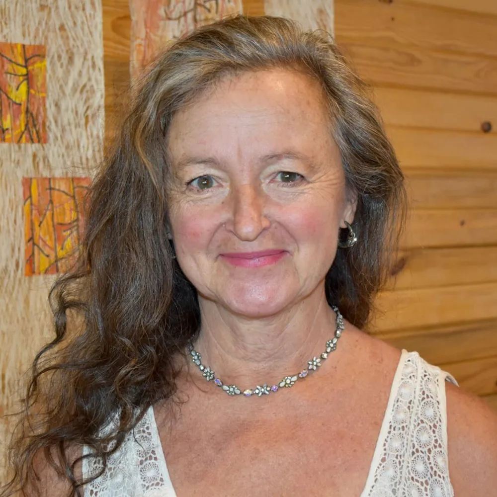Susan Nemcek smiling in a white top and beaded necklace, against a warm wood-slat wall.
