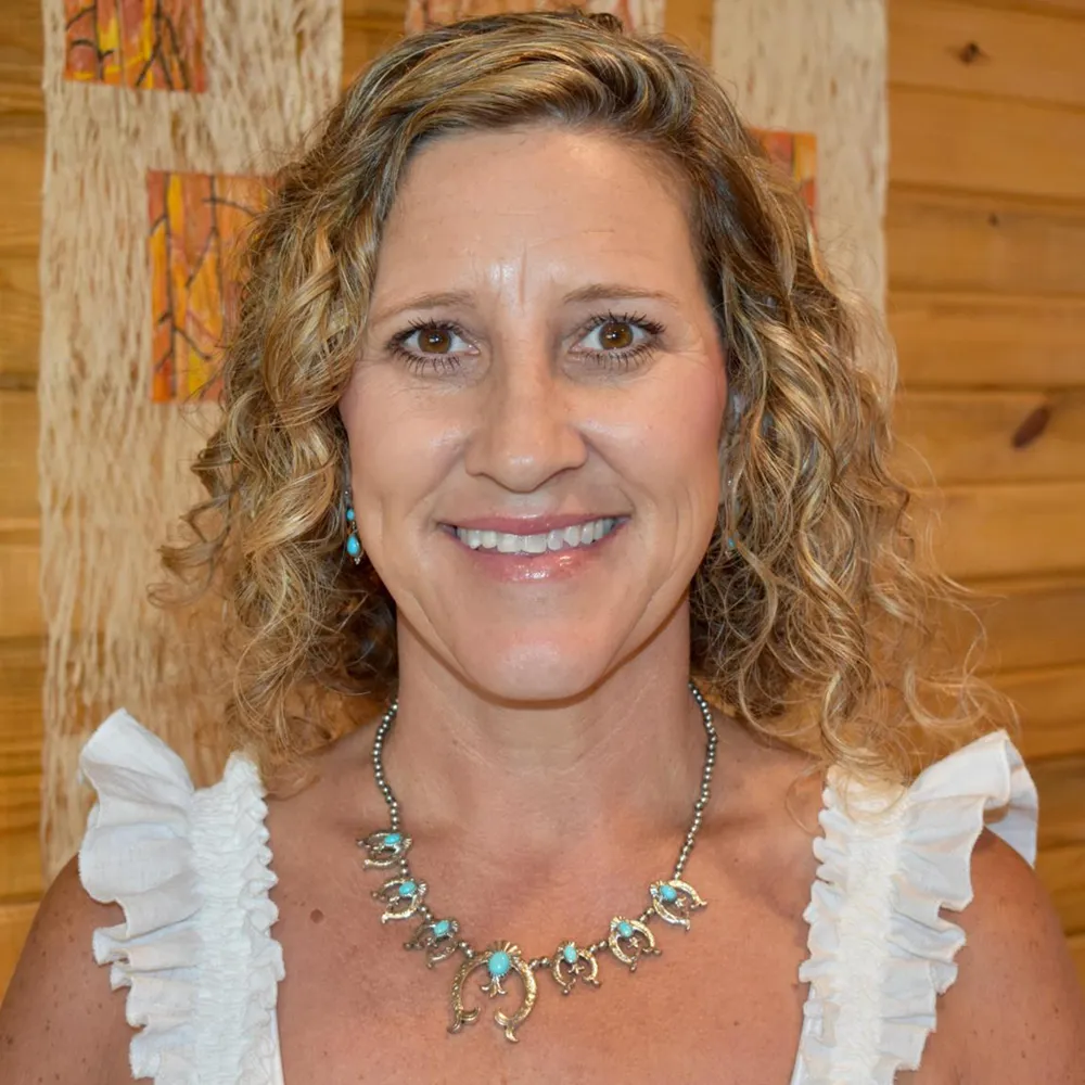 Vicki Viddal smiling warmly in a white blouse and turquoise necklace, against a warm wood-slat wall.